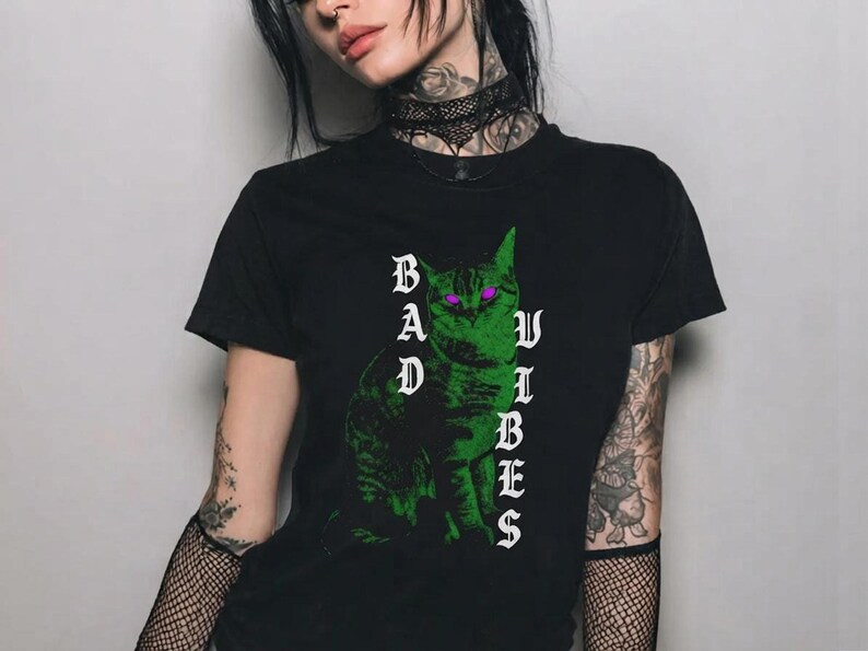 Close-up of a tattooed person wearing the black graphic t-shirt featuring a green cat with purple eyes.