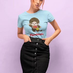 May include: Light blue t-shirt featuring a cartoon Monchhichi character with a red bib and yellow bag. The text "Monchhichi" is printed in red below the graphic. The shirt is paired with a black denim button-down skirt.