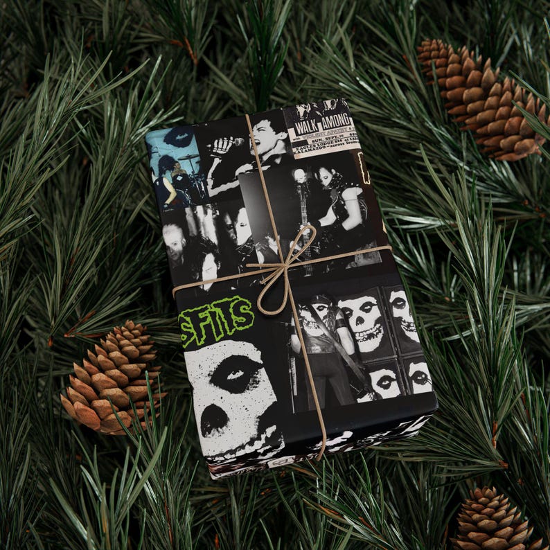 A gift wrapped in black and white Misfits-themed paper with twine, resting on green pine branches with pinecones.