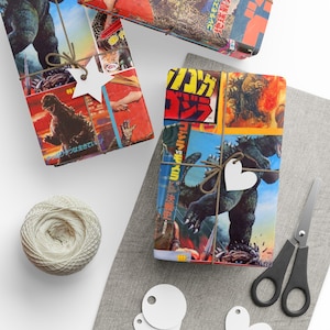 Two rectangular gifts wrapped in Godzilla-themed paper, one with a white star and the other with a white heart, on a gray mat with twine and scissors.