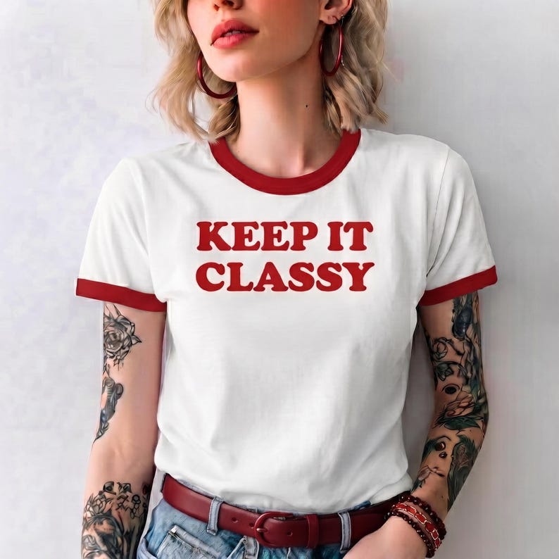 May include: A white ringer t-shirt with red trim on the collar and sleeve cuffs. The shirt features the red text "KEEP IT CLASSY" printed in a bold, sans-serif font across the center of the chest. The garment is worn with a dark red belt.
