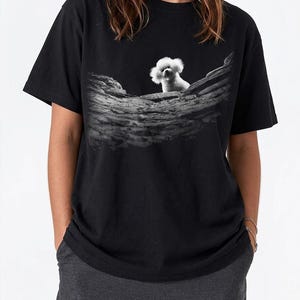 May include: A black short-sleeved t-shirt featuring a graphic print on the chest. The print displays a black and white image of a small, fluffy white dog peering over a textured, weathered wooden log or branch set against a dark background.