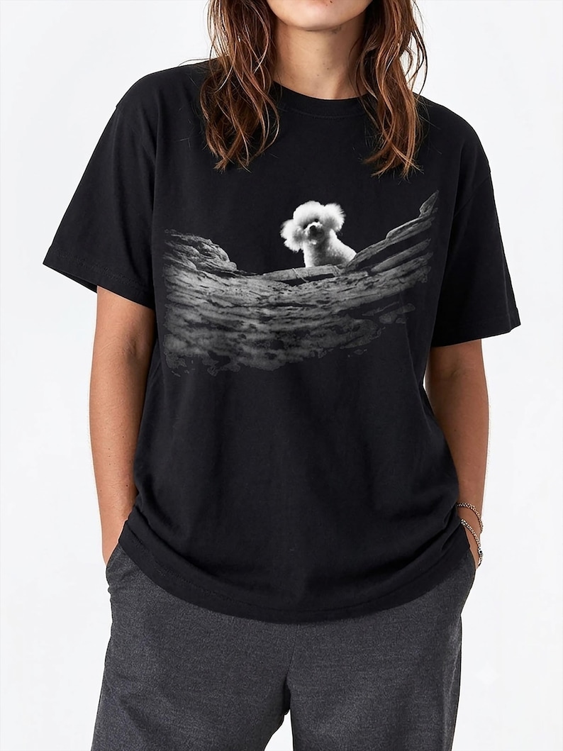 May include: A black short-sleeved t-shirt featuring a graphic print on the chest. The print displays a black and white image of a small, fluffy white dog peering over a textured, weathered wooden log or branch set against a dark background.