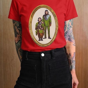 Könnte beinhalten: Ein leuchtend rotes T-Shirt mit kurzen Ärmeln und einem ovalen Grafikdruck in der Mitte. Der Druck zeigt eine Familie aus drei anthropomorphen Schimpansen in grüner und brauner Kleidung, eingefasst in einen goldenen Rahmen.