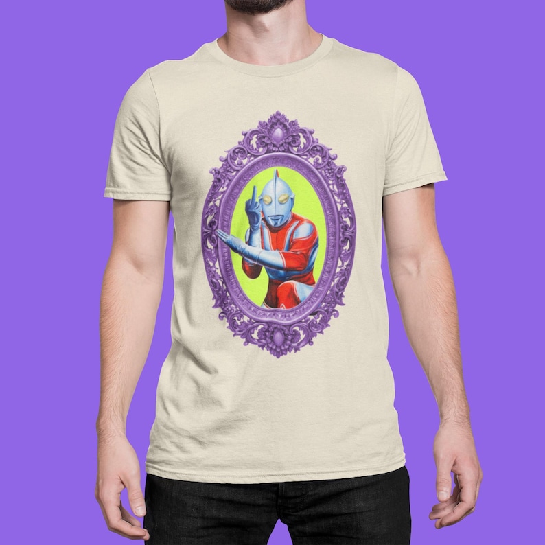 May include: Beige t-shirt with a framed illustration of a retro superhero figure in red, blue, and silver, giving the middle finger. The purple frame has ornate detailing, set against a lime green background.