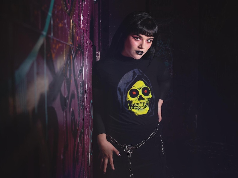 A person with dark hair and makeup wears a black long-sleeve shirt with a yellow skull in a blue hood, leaning against a graffiti-covered wall.