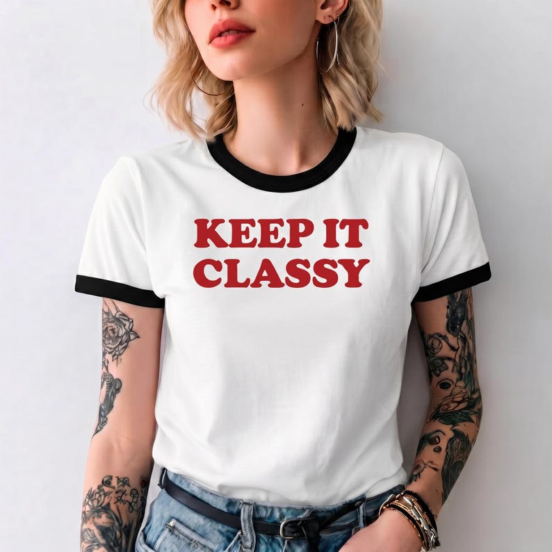 May include: A white ringer t-shirt with black trim on the collar and sleeve cuffs. The shirt features the text KEEP IT CLASSY printed in bold red capital letters across the center of the chest. The garment is made of a lightweight fabric.