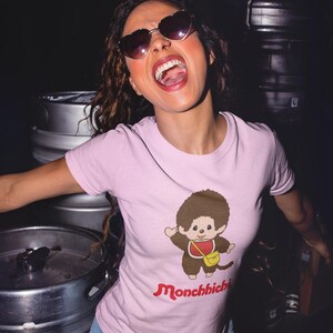 A woman wearing heart-shaped sunglasses and a pink t-shirt with a Monchhichi graphic, posing in front of metal barrels.
