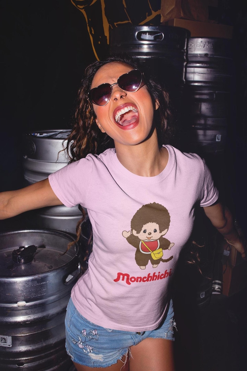 A woman wearing heart-shaped sunglasses and a pink t-shirt with a Monchhichi graphic, posing in front of metal barrels.