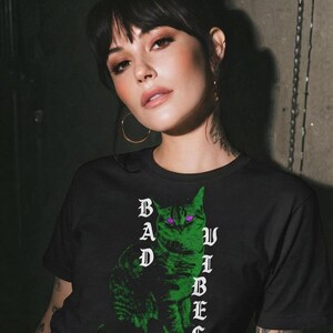 A woman with dark hair and hoop earrings poses in a black graphic t-shirt displaying a neon green cat with purple eyes.