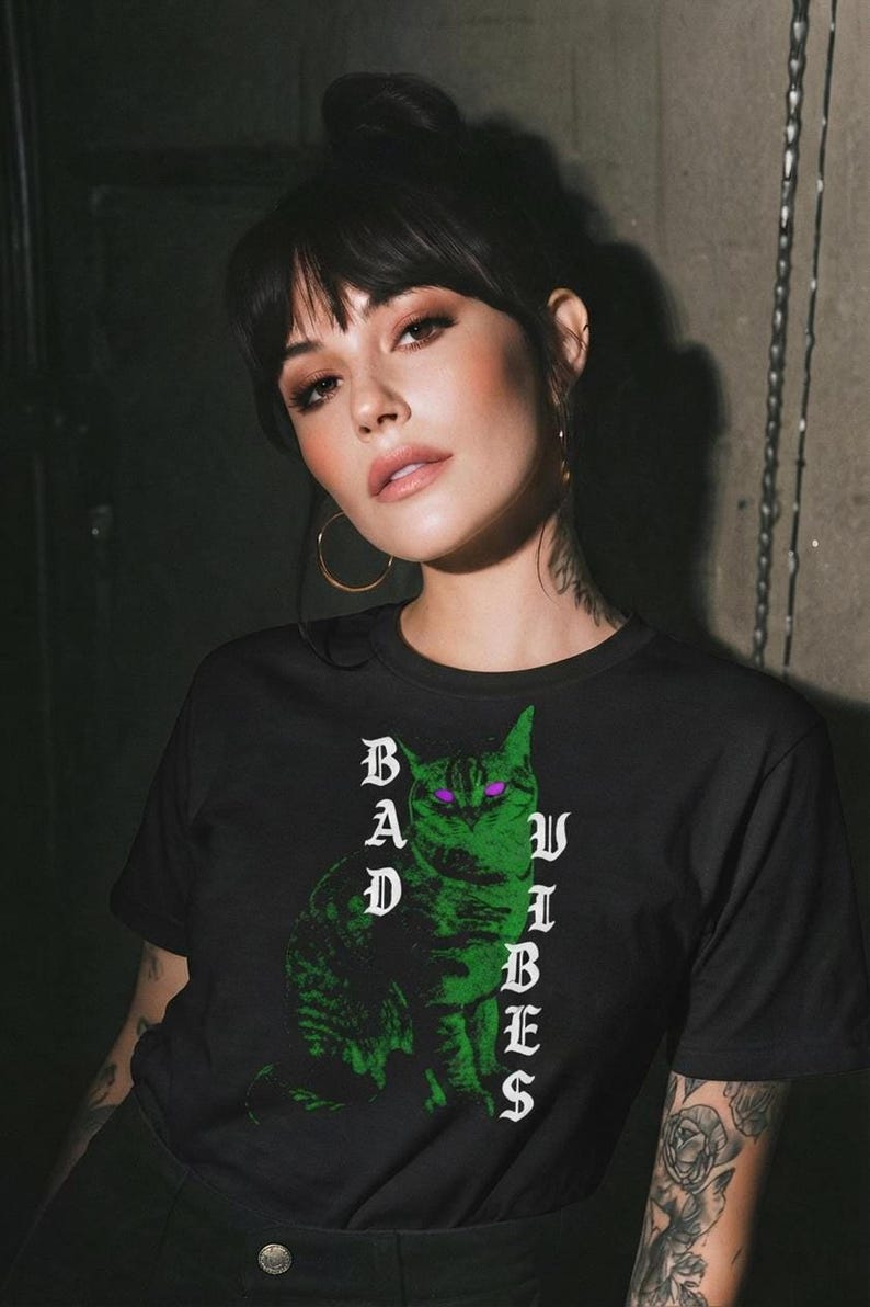 A woman with dark hair and hoop earrings poses in a black graphic t-shirt displaying a neon green cat with purple eyes.