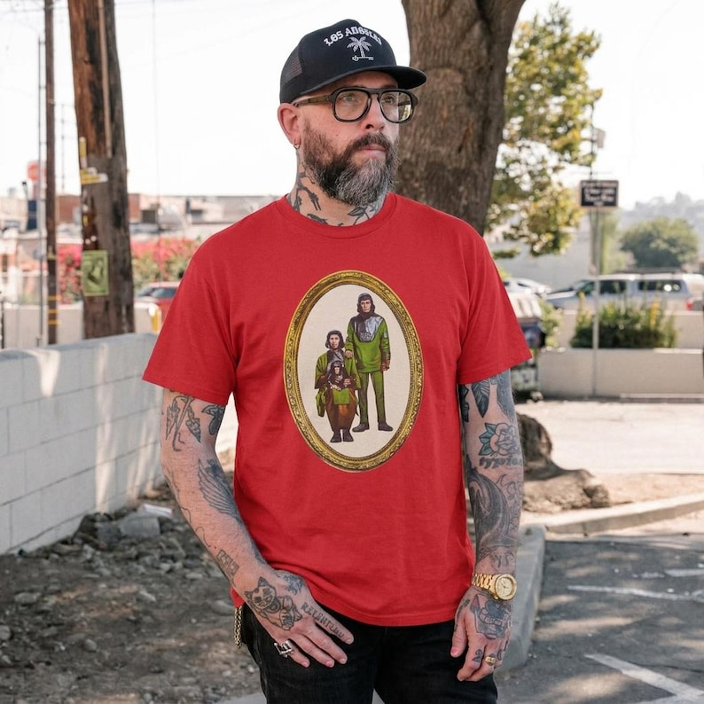 May include: A bright red short-sleeved t-shirt featuring a central oval graphic print of two anthropomorphic ape characters in green tunics. The design is enclosed within a detailed gold-colored frame, creating a portrait-style illustration on the chest.
