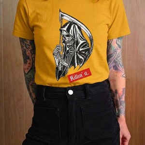A person with red hair wearing a mustard yellow t-shirt featuring a graphic of the grim reaper holding a scythe.