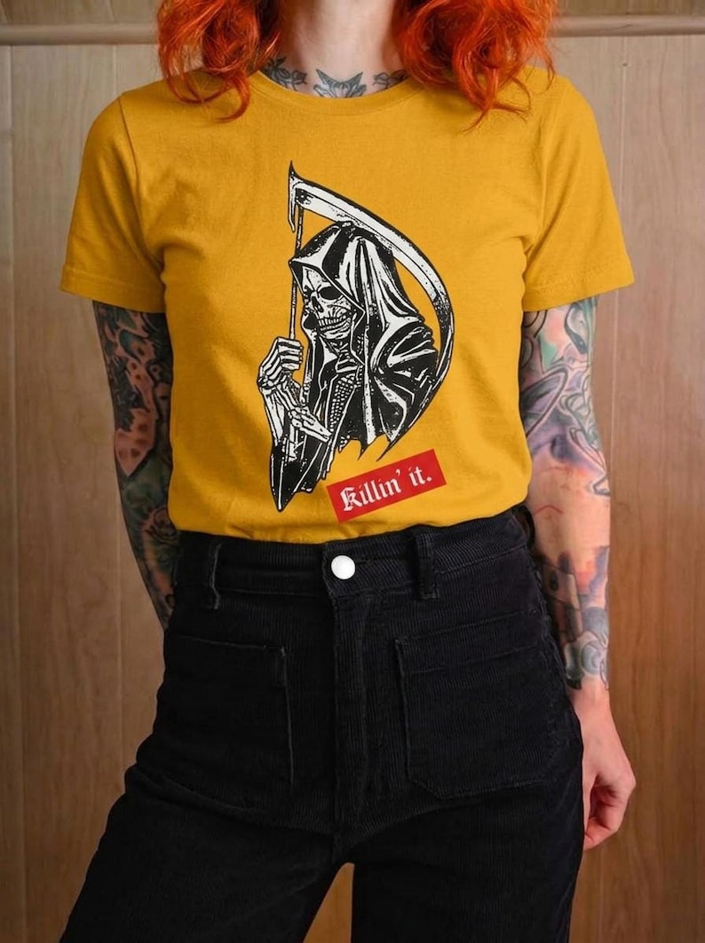 A person with red hair wearing a mustard yellow t-shirt featuring a graphic of the grim reaper holding a scythe.