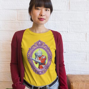May include: Yellow raglan t-shirt with a framed illustration of a superhero in red and silver. The shirt is worn with a burgundy cardigan and blue jeans. The image is set against a white brick wall.