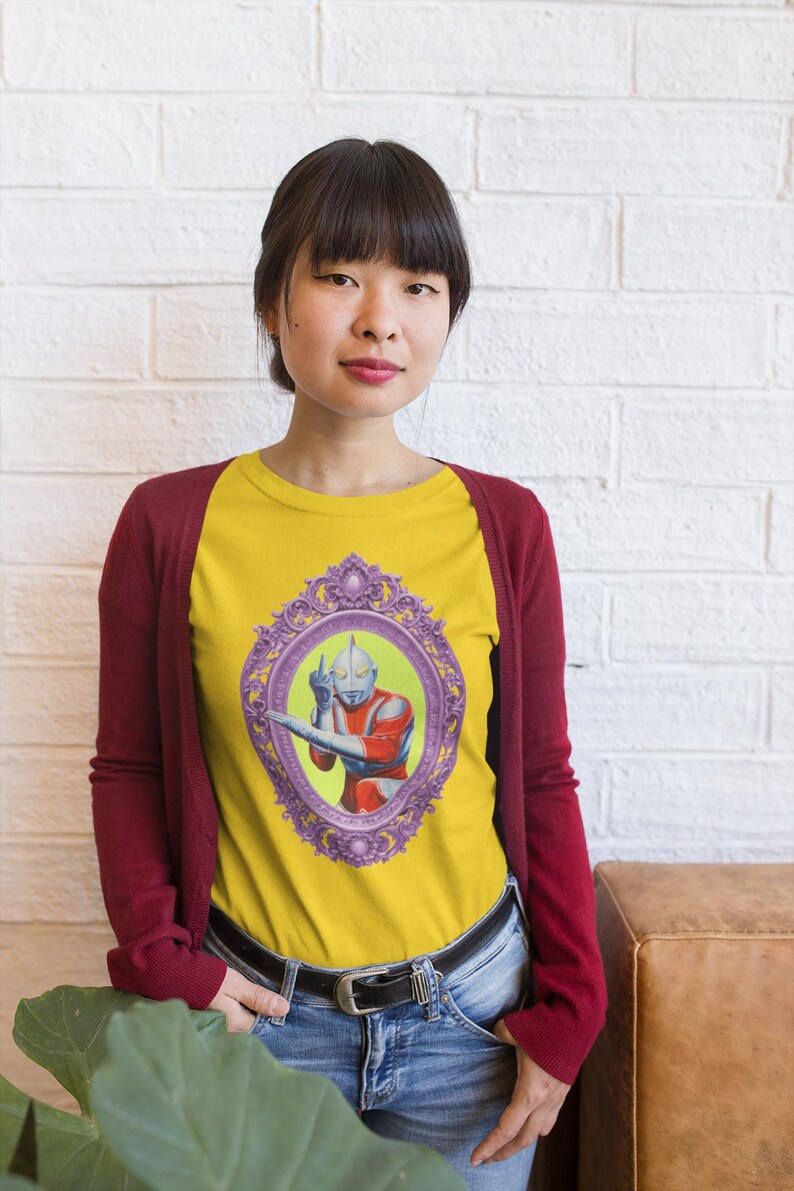 May include: Yellow raglan t-shirt with a framed illustration of a superhero in red and silver. The shirt is worn with a burgundy cardigan and blue jeans. The image is set against a white brick wall.