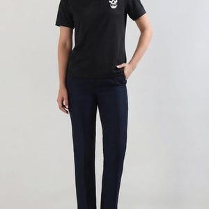 A person standing and wearing a black polo shirt featuring a white skull patch on the chest, paired with dark trousers.
