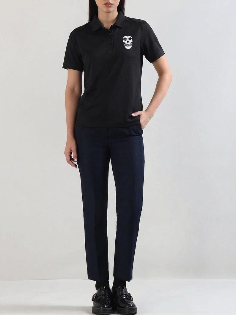 A person standing and wearing a black polo shirt featuring a white skull patch on the chest, paired with dark trousers.