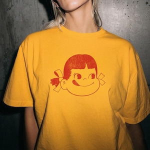 A close-up of a person wearing a mustard yellow t-shirt with a red graphic of a girl's face with pigtails and a winking expression.