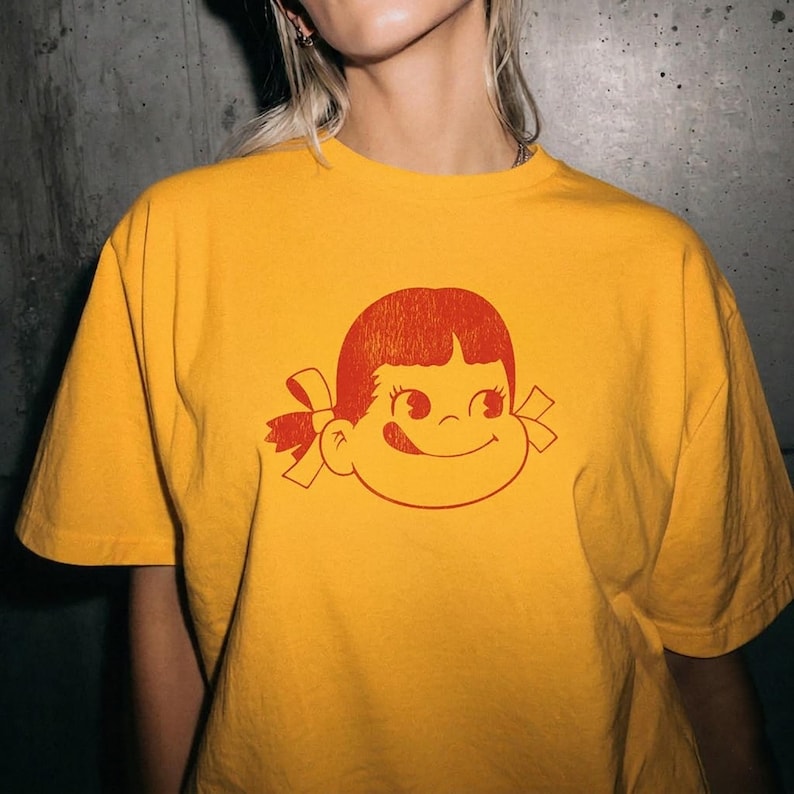 A close-up of a person wearing a mustard yellow t-shirt with a red graphic of a girl's face with pigtails and a winking expression.