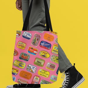 A person wearing a white shirt, gray pants, and black sneakers holds a pink tote bag with sardine can labels, against a yellow background.