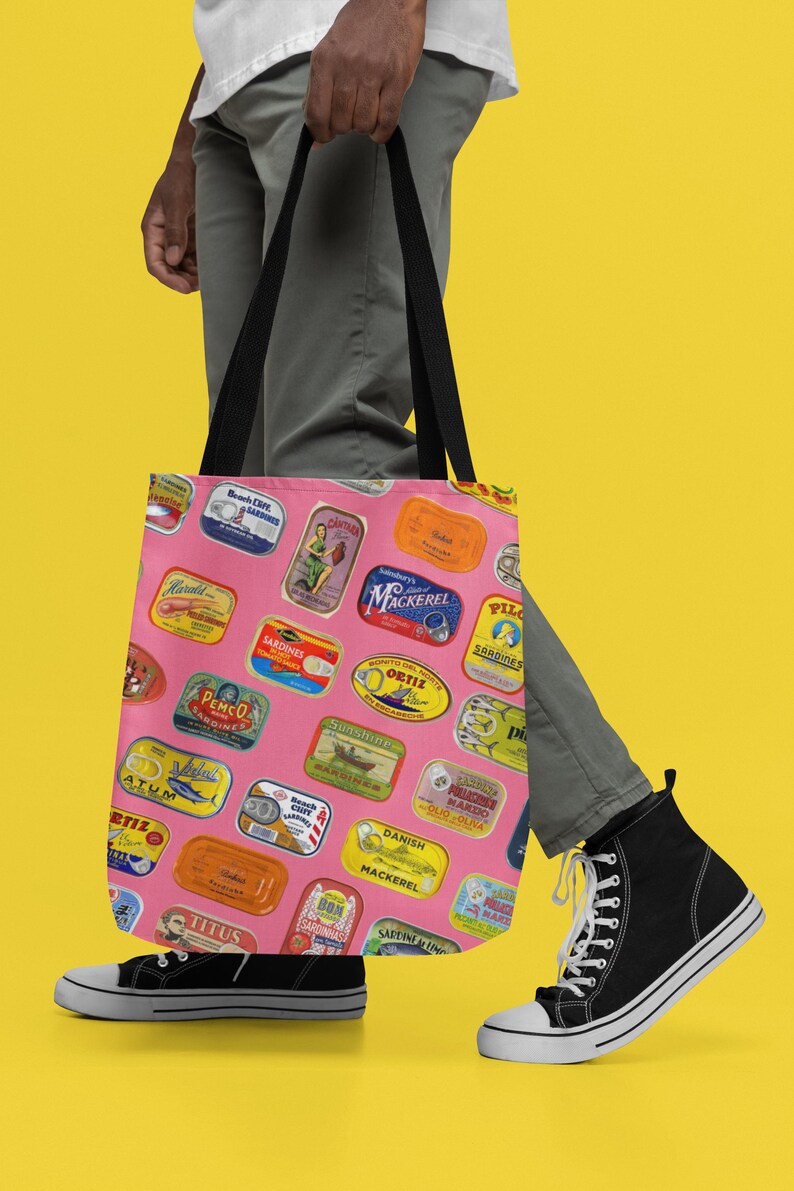 A person wearing a white shirt, gray pants, and black sneakers holds a pink tote bag with sardine can labels, against a yellow background.