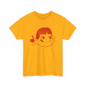 A flat lay of a mustard yellow t-shirt with a red graphic of a girl's face with pigtails and a winking expression.