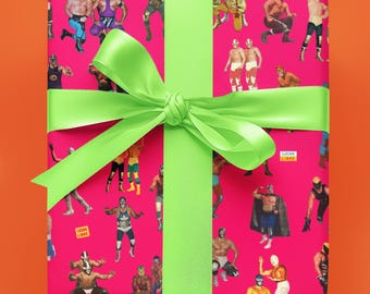 May include: A pink gift box with a green ribbon. The box is wrapped in wrapping paper with a pattern of wrestlers wearing masks. The text "LUCHA LIBRE" is printed on the wrapping paper.
