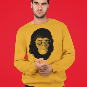 K&ouml;nnte beinhalten: Senfgelbes Sweatshirt mit einem schwarz-wei&szlig;en Grafik eines Affen mit Sonnenbrille. Das Gesicht des Affen ist mittig auf der Vorderseite des lang&auml;rmeligen Rundhalsausschnitts. Der Hintergrund ist einfarbig rot.