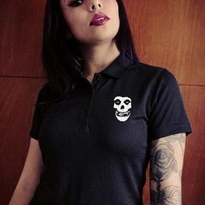 A person with dark hair and a rose tattoo on their arm wearing a black polo shirt with a white skull embroidery on the chest.