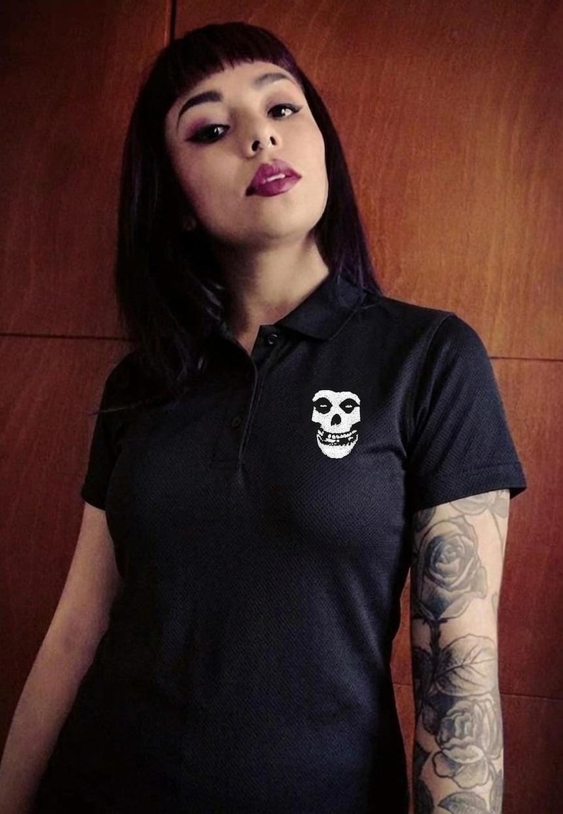 A person with dark hair and a rose tattoo on their arm wearing a black polo shirt with a white skull embroidery on the chest.