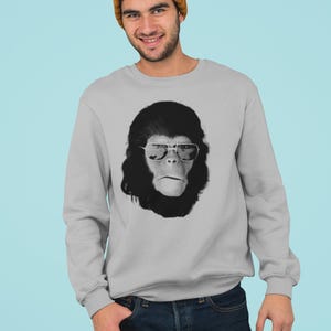 K&ouml;nnte beinhalten: Ein hellgraues Sweatshirt mit einem Schwarz-Wei&szlig;-Bild eines Affen mit Sonnenbrille. Die Person auf dem Bild tr&auml;gt eine senfgelbe M&uuml;tze und eine blaue Jeans. Der Hintergrund ist hellblau.