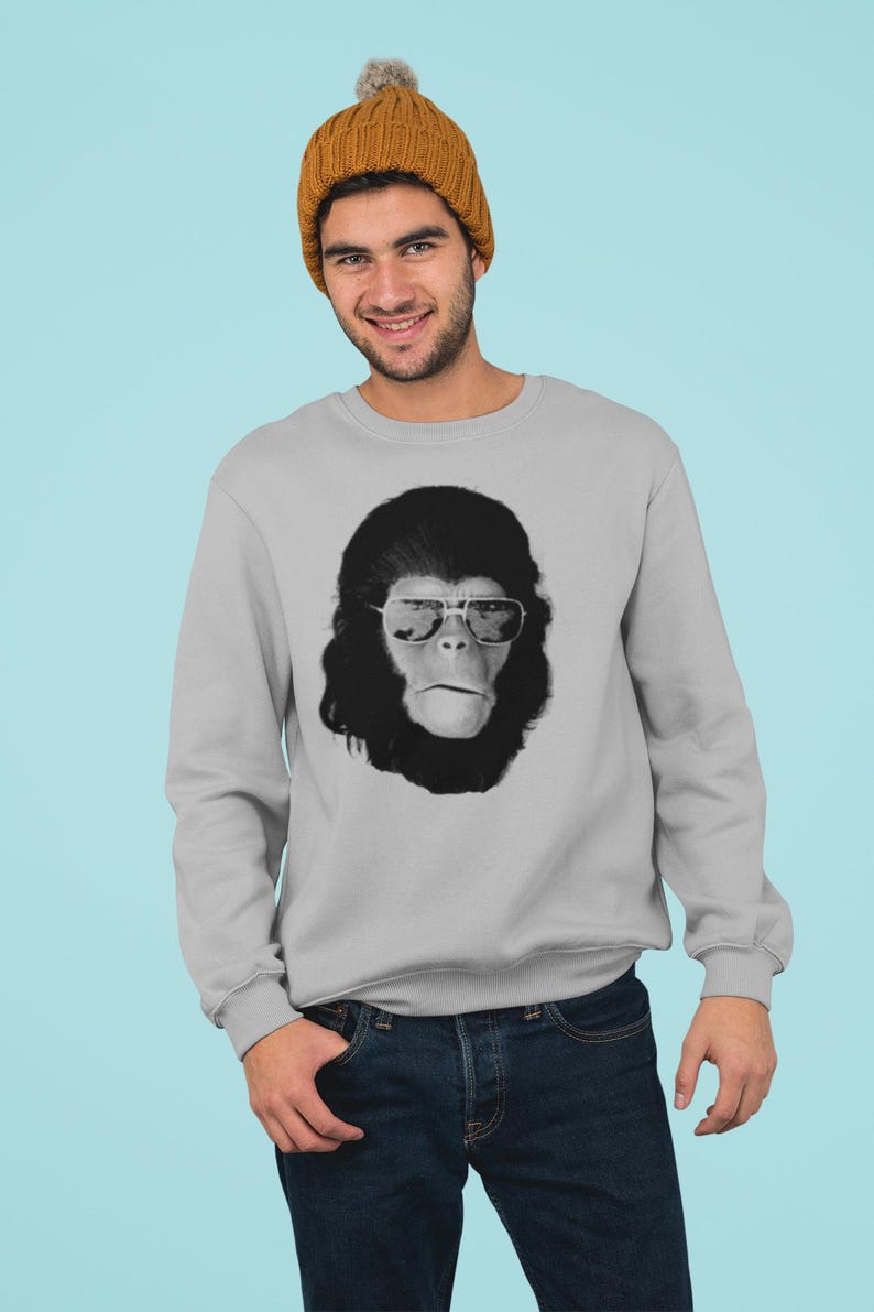 K&ouml;nnte beinhalten: Ein hellgraues Sweatshirt mit einem Schwarz-Wei&szlig;-Bild eines Affen mit Sonnenbrille. Die Person auf dem Bild tr&auml;gt eine senfgelbe M&uuml;tze und eine blaue Jeans. Der Hintergrund ist hellblau.