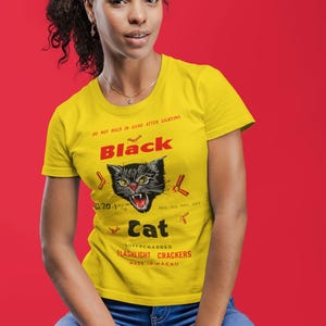 May include: A yellow t-shirt featuring a black cat graphic and the words "Black Cat Supercharged Flashlight Crackers". The shirt also has the text "Do not hold in hand after lighting". The model is wearing blue jeans.