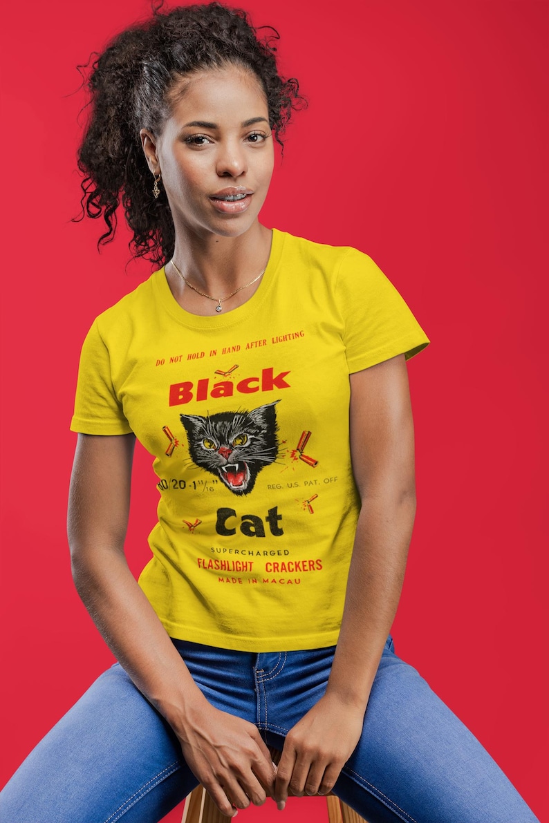 May include: A yellow t-shirt featuring a black cat graphic and the words "Black Cat Supercharged Flashlight Crackers". The shirt also has the text "Do not hold in hand after lighting". The model is wearing blue jeans.