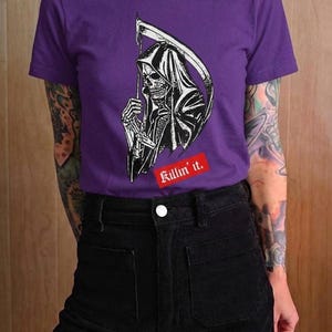 A person with bright red hair wearing a purple t-shirt with a white grim reaper illustration and red text.