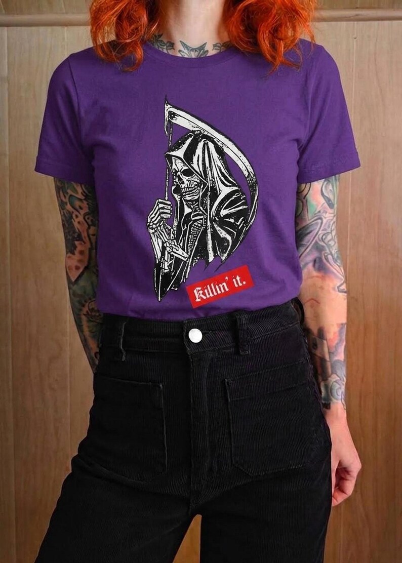 A person with bright red hair wearing a purple t-shirt with a white grim reaper illustration and red text.