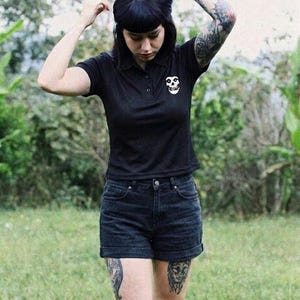 A tattooed person with dark bangs wearing a black polo shirt with a white skull patch and black shorts outdoors in a grassy area.
