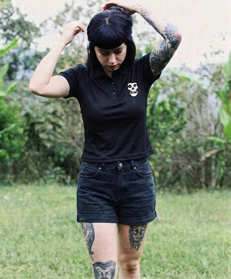 A tattooed person with dark bangs wearing a black polo shirt with a white skull patch and black shorts outdoors in a grassy area.