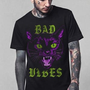 May include: A black short-sleeved t-shirt featuring a graphic print of a snarling cat face in shades of purple and green. The text "BAD" is printed in green above the cat, and the word "VIBES" is printed in green below the cat illustration.