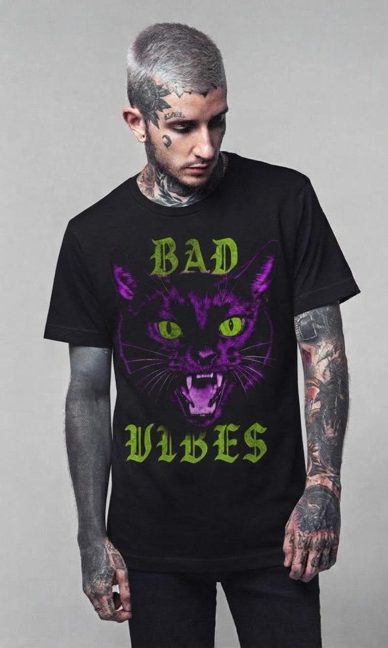 May include: A black short-sleeved t-shirt featuring a graphic print of a snarling cat face in shades of purple and green. The text "BAD" is printed in green above the cat, and the word "VIBES" is printed in green below the cat illustration.