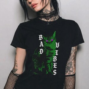 A tattooed person wearing a black t-shirt with a green cat graphic and mesh sleeves underneath.