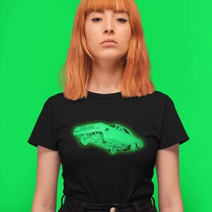 May include: A black short-sleeve t-shirt featuring a glowing neon green graphic of a classic sedan car on the chest. The shirt has a crew neckline and is paired with a black skirt that has a button-down front closure and visible metal buttons.