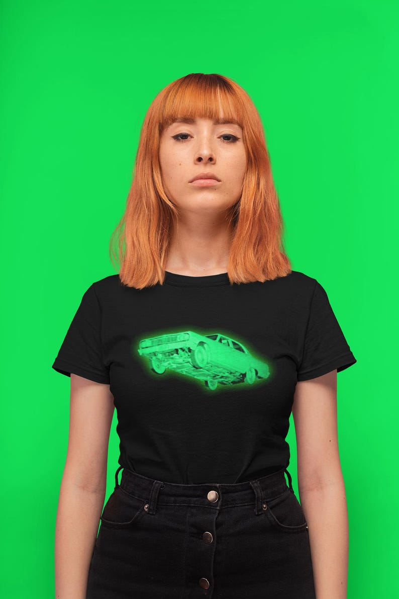 May include: A black short-sleeve t-shirt featuring a glowing neon green graphic of a classic sedan car on the chest. The shirt has a crew neckline and is paired with a black skirt that has a button-down front closure and visible metal buttons.