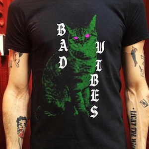 A tattooed man wearing a black t-shirt featuring a green cat graphic and the text Bad Vibes in white gothic lettering.