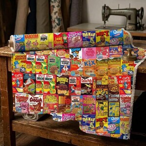 May include: A roll of fabric featuring a repeating grid pattern of vintage cereal box designs. The colorful print includes various iconic breakfast cereal brands, cartoon mascots, and product logos arranged in a structured, multi-colored collage layout.