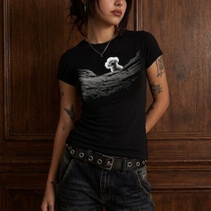 May include: A black short-sleeved t-shirt featuring a graphic print of a small white fluffy dog peeking over a textured, rocky, grey landscape. The shirt has a crew neckline and a fitted silhouette, paired with dark blue denim jeans and a black belt.