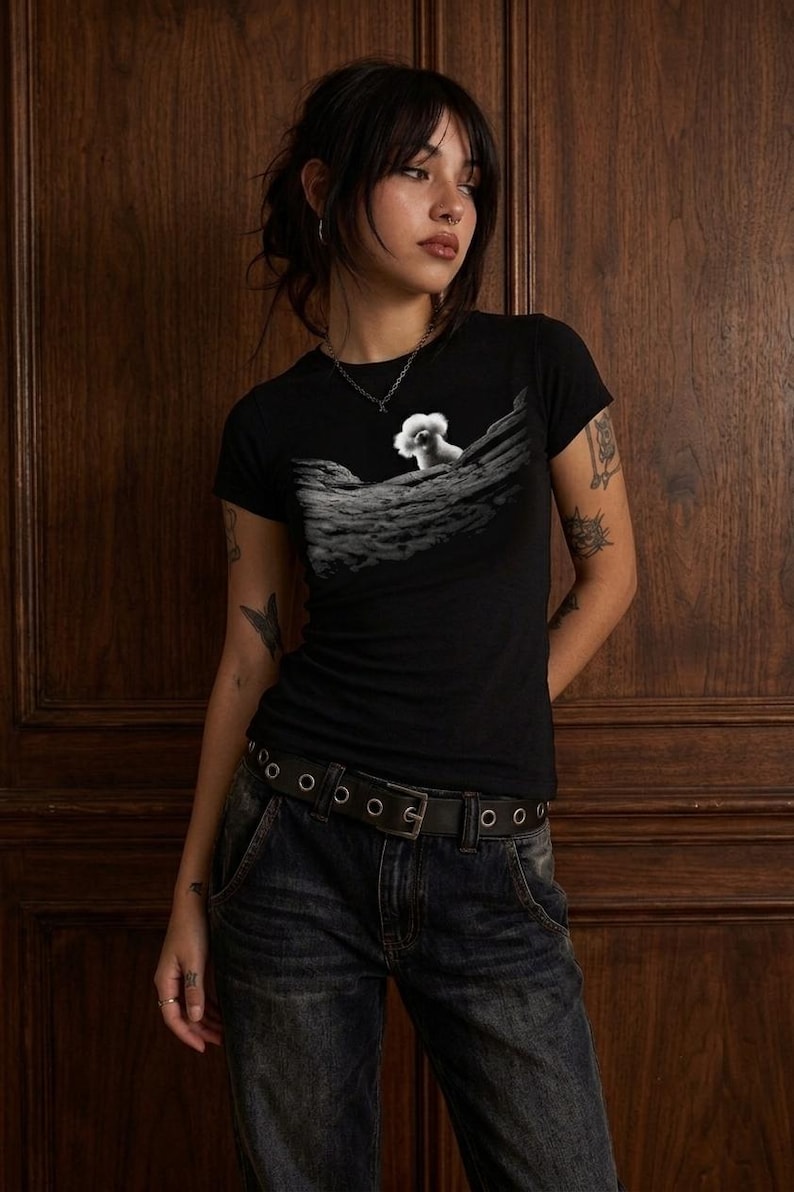 May include: A black short-sleeved t-shirt featuring a graphic print of a small white fluffy dog peeking over a textured, rocky, grey landscape. The shirt has a crew neckline and a fitted silhouette, paired with dark blue denim jeans and a black belt.
