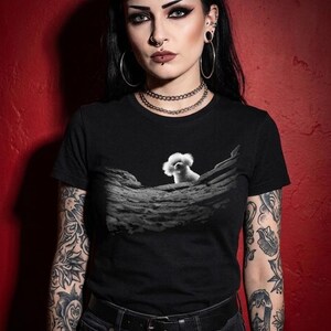 May include: A black short-sleeved t-shirt featuring a graphic print on the chest. The print displays a small white fluffy dog peeking over a textured, rocky, or moon-like landscape rendered in shades of grey and black against the solid black fabric.