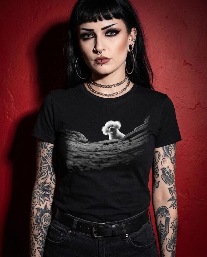 May include: A black short-sleeved t-shirt featuring a graphic print on the chest. The print displays a small white fluffy dog peeking over a textured, rocky, or moon-like landscape rendered in shades of grey and black against the solid black fabric.